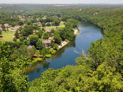 [A crescent-shaped body of water is rimmed by lush amounts of green-leaved trees. Some houses sit near the water's edge on the north side of the lake.]